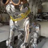 Mirror-finish dog sculpture with copper-toned glasses and a yellow chain, viewed at an angle in the workshop