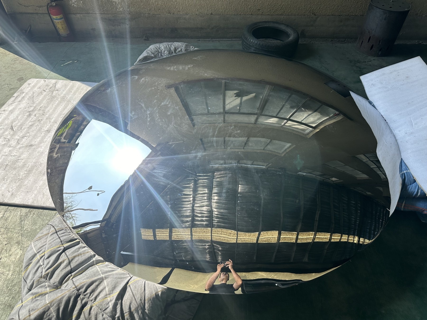 Angled workshop view of a convex stainless steel mirror disc showing its perimeter and reflected roof